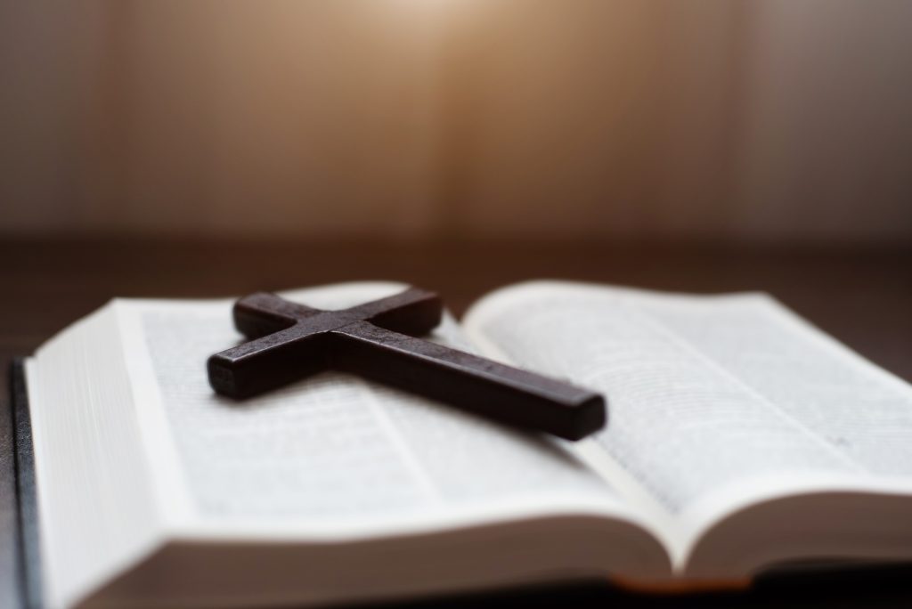 Close up of an open bible with a cross for morning devotion on a wooden table with window lights.